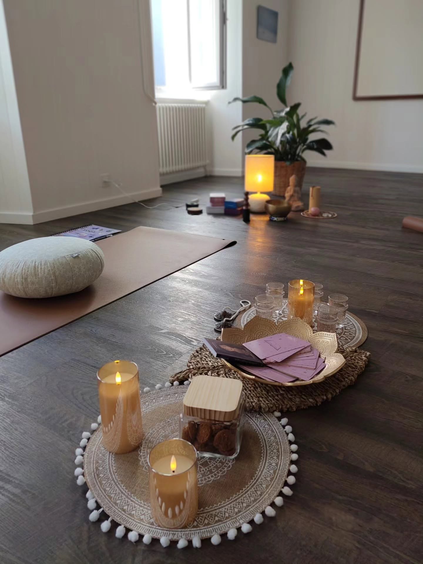 Un coin méditation du studio : un tapis déroulé, un coussin rond, des bougies allumées et un plateau en fibres naturelles sur un parquet sombre.